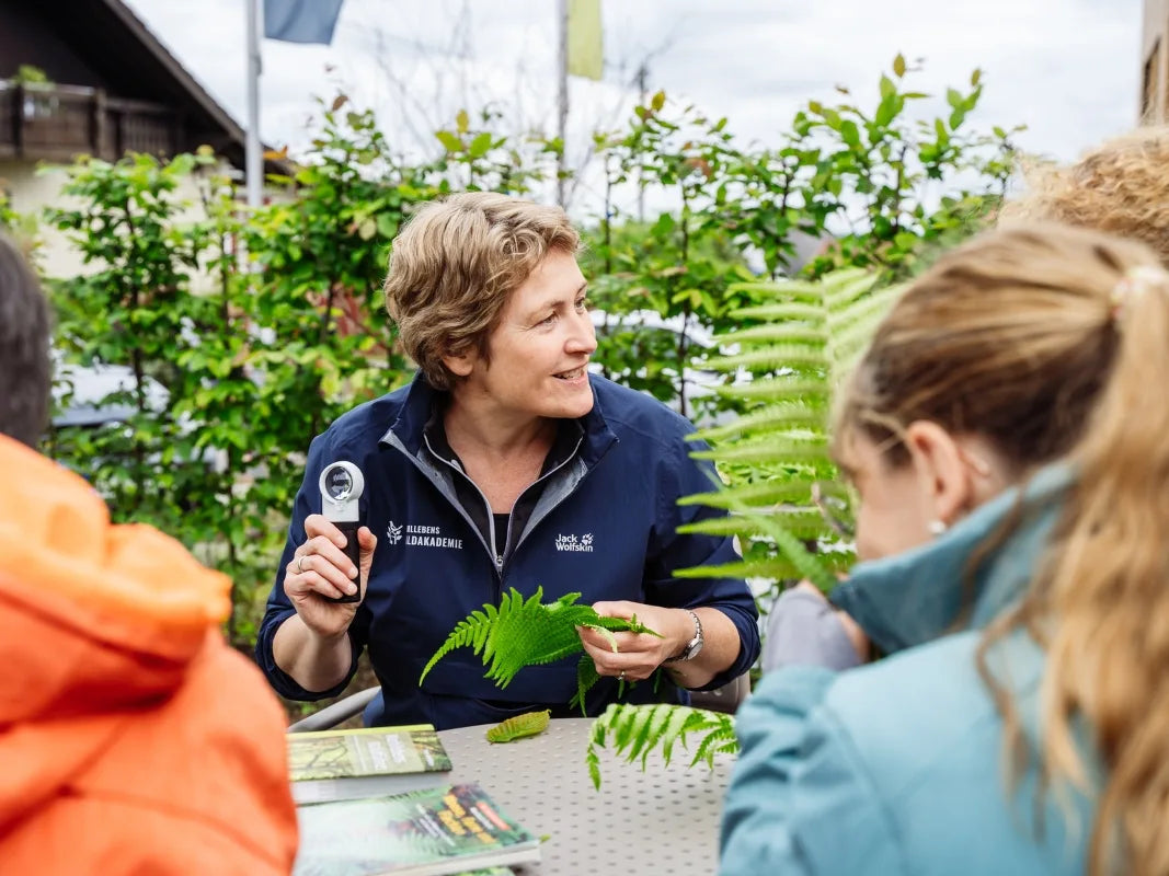 Eine Frau von Wohllebens Waldakademie leitet ein Pflanzenwissenschaft-Seminar im Freien und bringt den Kindern etwas über Farne bei, während sie eine Lupe und ein Farnblatt am Tisch hält.