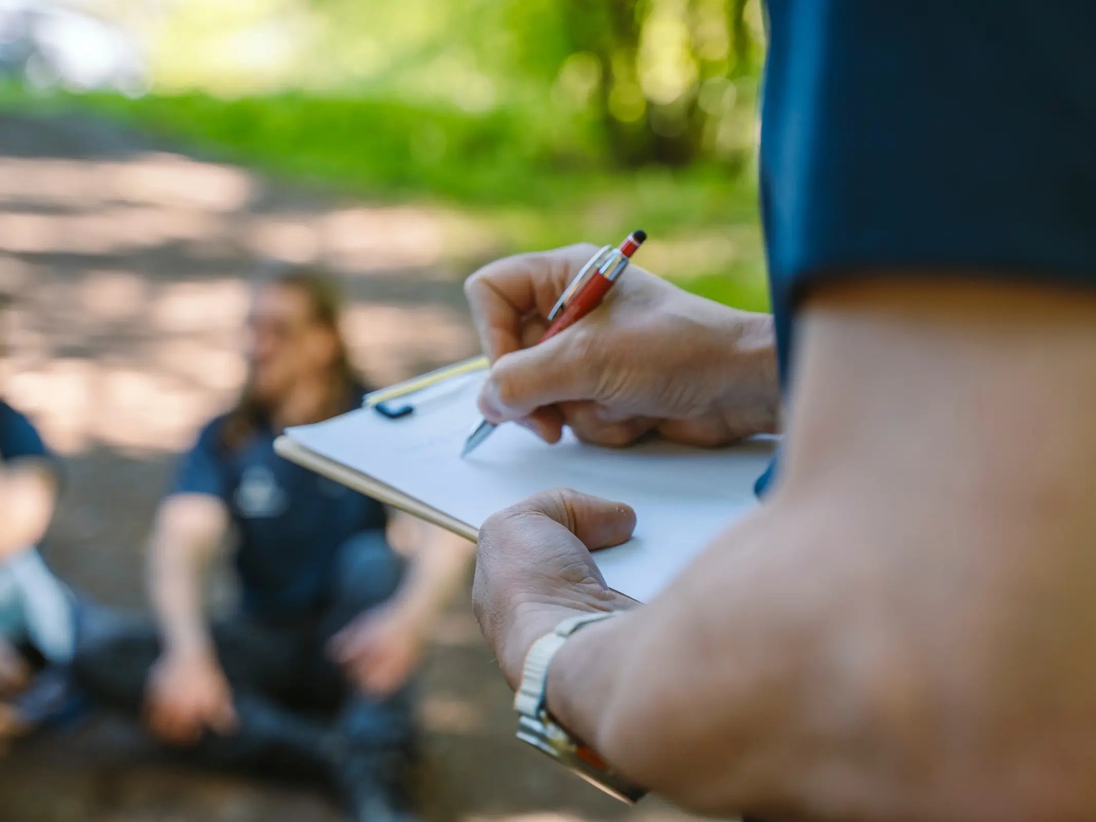 Eine Person schreibt im Freien auf einem Klemmbrett, möglicherweise um Erkenntnisse aus einem Kurs der Wohllebens Waldakademie zu dokumentieren, während im unscharfen Hintergrund Menschen sitzen.