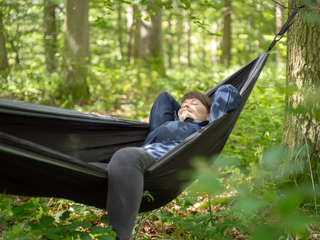 Eine Person entspannt sich in einer schwarzen Hängematte im grünen Wald, genießt ein echtes Eifelwald-Erlebnis und verbringt die Nacht unter dem Sternenhimmel bei Wohllebens Waldakademie Nacht im Wald.