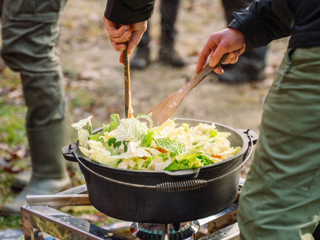 Das Kochen von Blattgemüse in einem großen schwarzen Topf über einem Gaskocher, dieses Outdoor-Erlebnis mit Wohllebens Waldakademie bringt frische Aromen zum Leben, während Sie eine Nacht im Wald unter freiem Himmel übernachten.