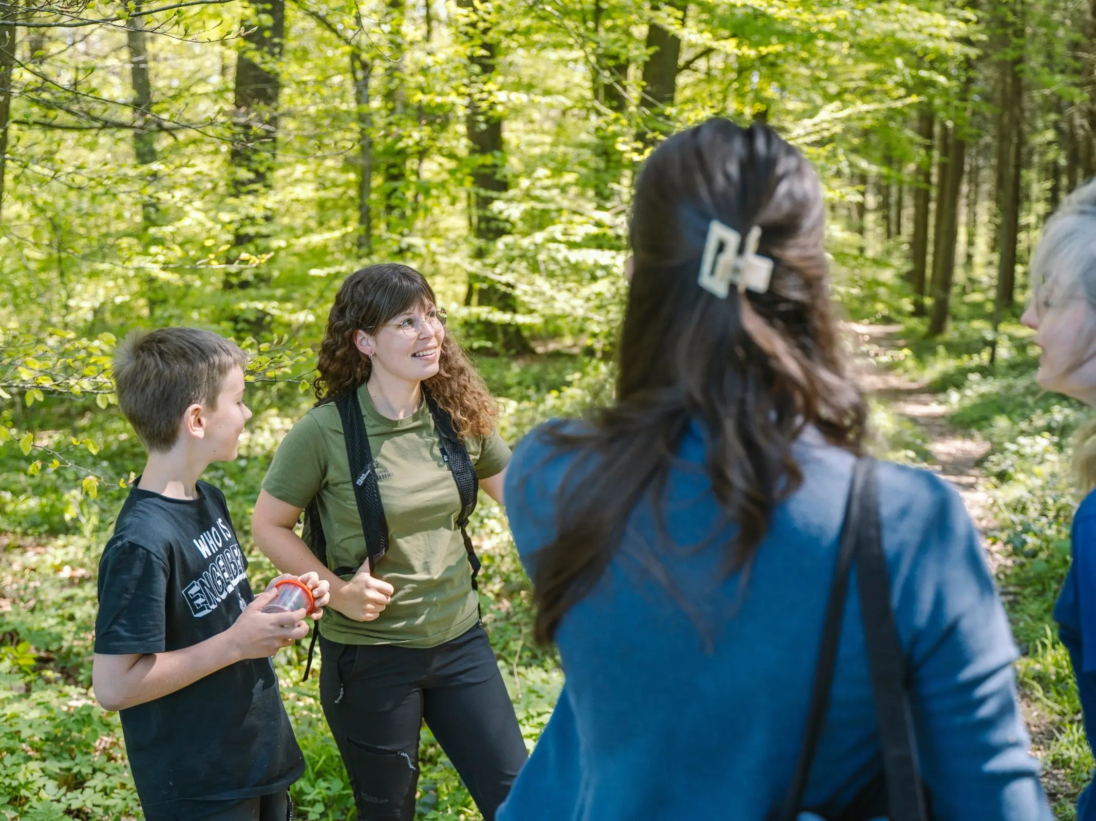 Eine Gruppe genießt die Familienwanderung Eifel mit Wohllebens Waldakademie und unterhält sich auf einem sonnenbeschienenen Waldweg, der von grünen Bäumen umgeben ist.