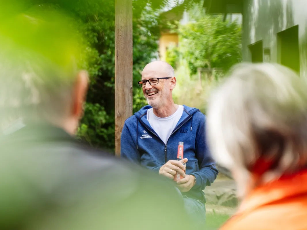 Ein Mann mit Brille lächelt, während er im Freien sitzt und sich mit anderen in einem Garten während eines veganen BBQs von Wohllebens Waldakademie unterhält.