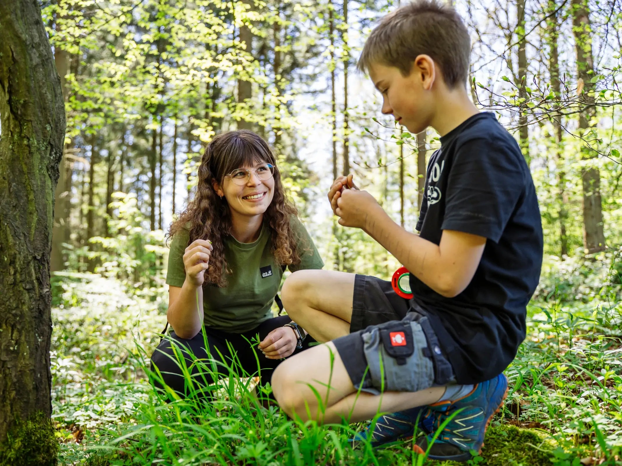 Eine Frau und ein Junge, die möglicherweise an der Wohllebens Waldakademie Juniorförster-Ausbildung oder am Waldkurs 7-10 Jahre teilnehmen, lächeln, während sie zwischen dem sonnenbeschienenen Laub des Waldes hocken und gemeinsam Pflanzen untersuchen.