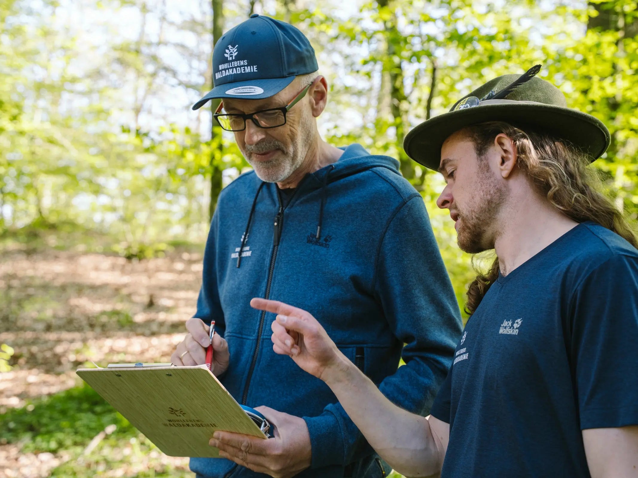 Zwei Personen mit Hüten diskutieren und schauen auf ein Klemmbrett, während sie in einem sonnenbeschienenen Wald Wissen von Wohllebens Waldakademie weitergeben.