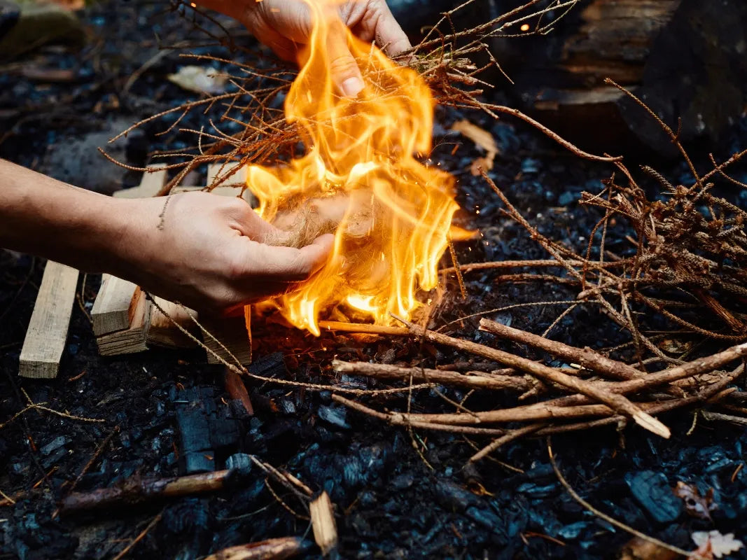 Hände, die ein kleines Lagerfeuer mit Zweigen auf verkohltem Boden im Freien entzünden - ein echter Wohllebens Waldakademie Survival Eifel-Moment, perfekt für jedes Abenteuer Wald.