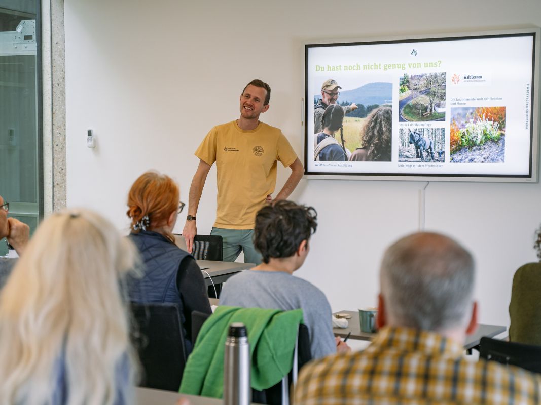 Ein Mann präsentiert einer kleinen Gruppe in einem Klassenzimmer eine Diashow mit Naturfotos und vermittelt Erkenntnisse aus der Wohllebens Waldakademie und dem Konzept Erlebnis Wald.