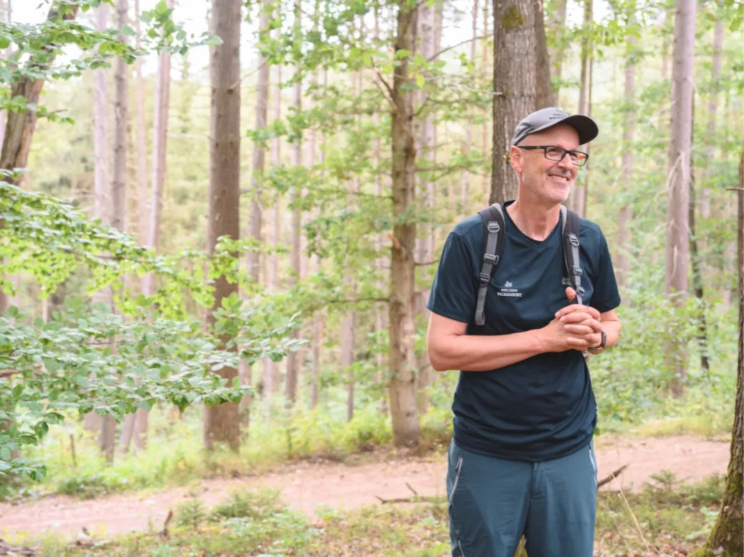 Ein Mann mit Brille und Rucksack lächelt in einem Buchenwälder Wald und genießt den strahlenden Tag bei einer Wanderung mit Wohllebens Waldakademie.