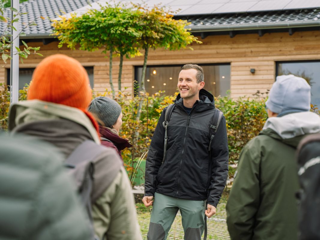 Ein lächelnder Mann in Outdoor-Ausrüstung teilt Weihnachtsfeier Ideen für eine Winterwanderung Gruppe vor einem Holzgebäude, das Wohllebens Waldakademie repräsentiert.