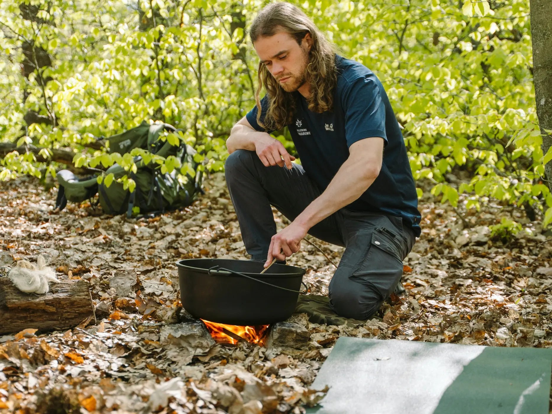 Eine Person kocht über einem kleinen Lagerfeuer im Wald und rührt einen Topf um - ein authentisches Erlebnis der Wohllebens Waldakademie und die perfekte Art, eine Nacht unter freiem Himmel zu genießen.