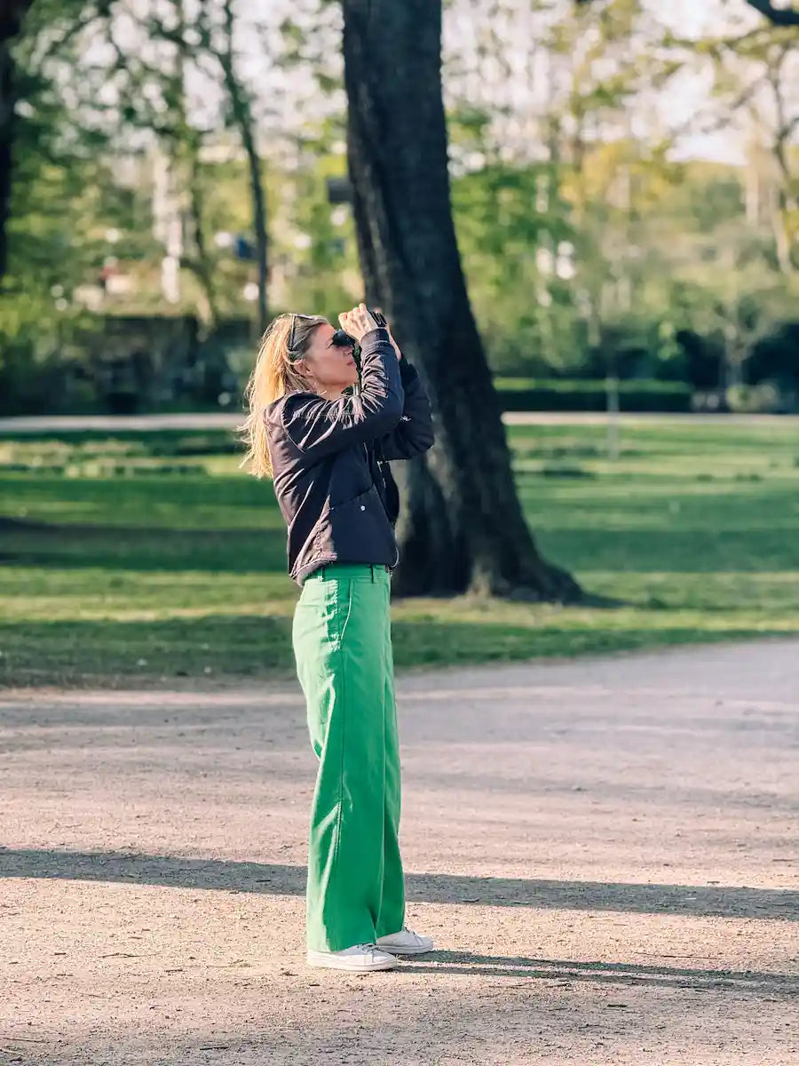 Eine Frau in grüner Hose und Sonnenbrille sucht mit einem Fernglas einen sonnigen Park ab und genießt die Vogelbeobachtung - vielleicht inspiriert von den Expertentipps der Wohllebens Waldakademie zur Beobachtung von Vögeln in Köln.