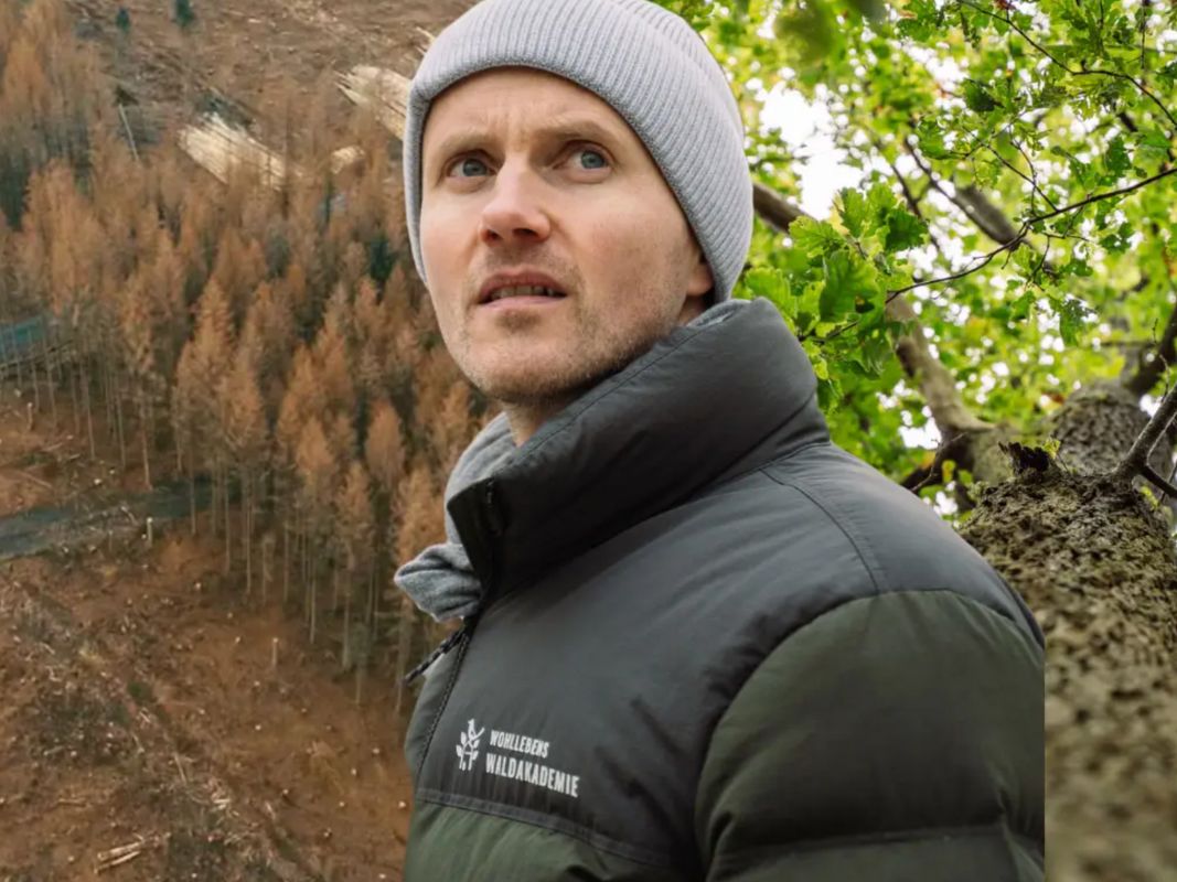 Ein Mann mit grauer Mütze und Mantel steht im Freien in der Nähe von Bäumen und macht sich Gedanken über die Gesundheit der Wälder und die Auswirkungen des Klimawandels, wie von der Wohllebens Waldakademie hervorgehoben.