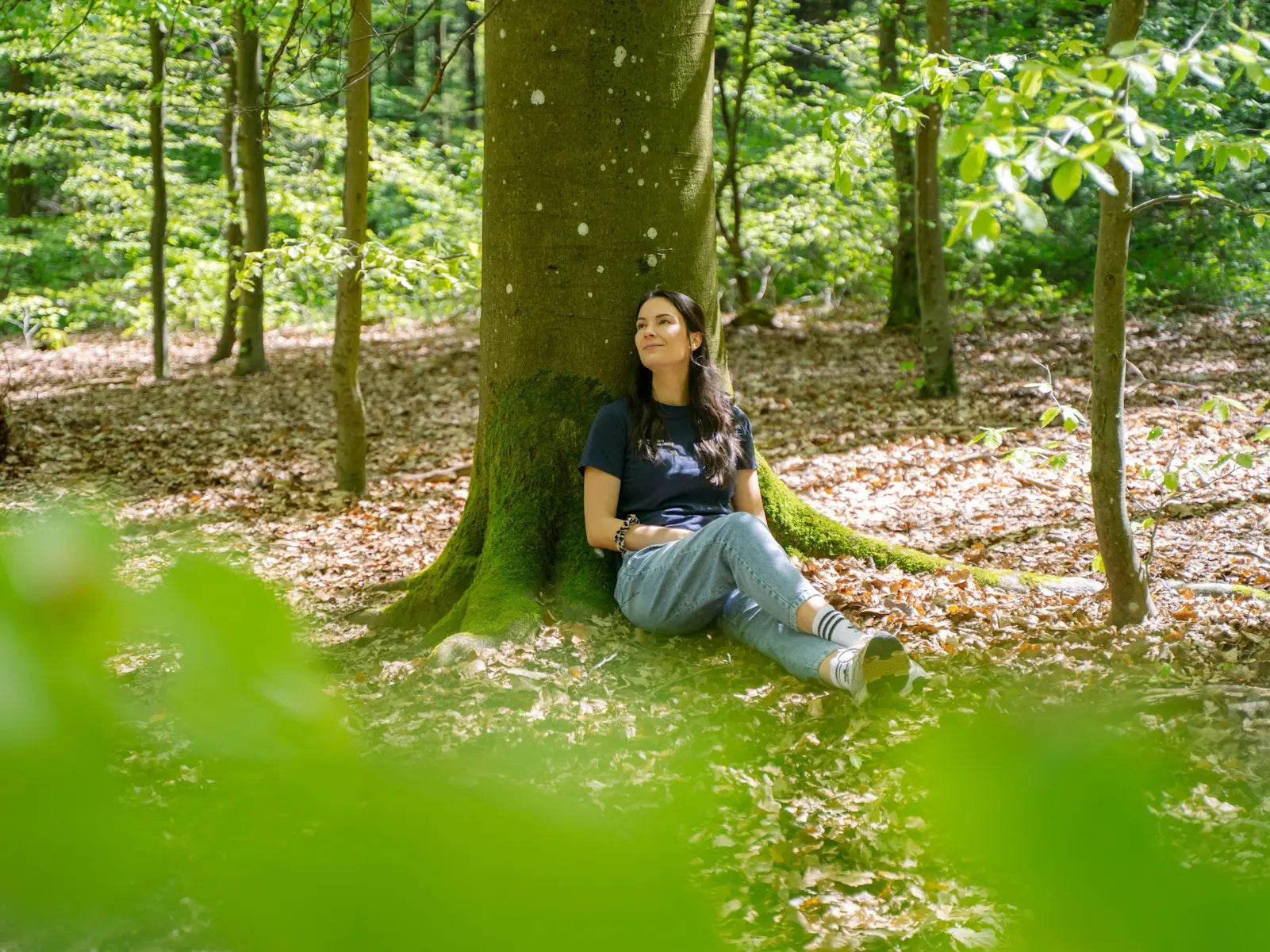 Eine Frau entspannt sich an einem Baumstamm und genießt Waldbaden Eifel von Wohllebens Waldakademie in einem sonnenbeschienenen Wald, um ihre Sinne zu entspannen und die Entschleunigung zu genießen.