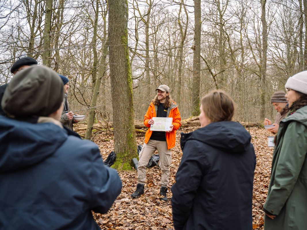 Ein Führer in einer orangefarbenen Jacke von Wohllebens Waldakademie hält eine Karte in der Hand und spricht mit einer Winterwanderungsgruppe in einem grünen Wald - eine tolle Inspiration für Weihnachtsfeier-Ideen.