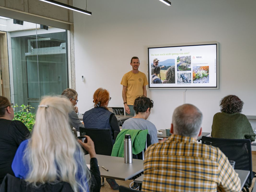 Ein Referent der Wohllebens Waldakademie spricht zu einer kleinen Gruppe in einem Klassenzimmer, zeigt Naturfotos von Wäldern und diskutiert über Umweltkrisen und ihre Auswirkungen auf unsere lebenswichtigen Ressourcen.