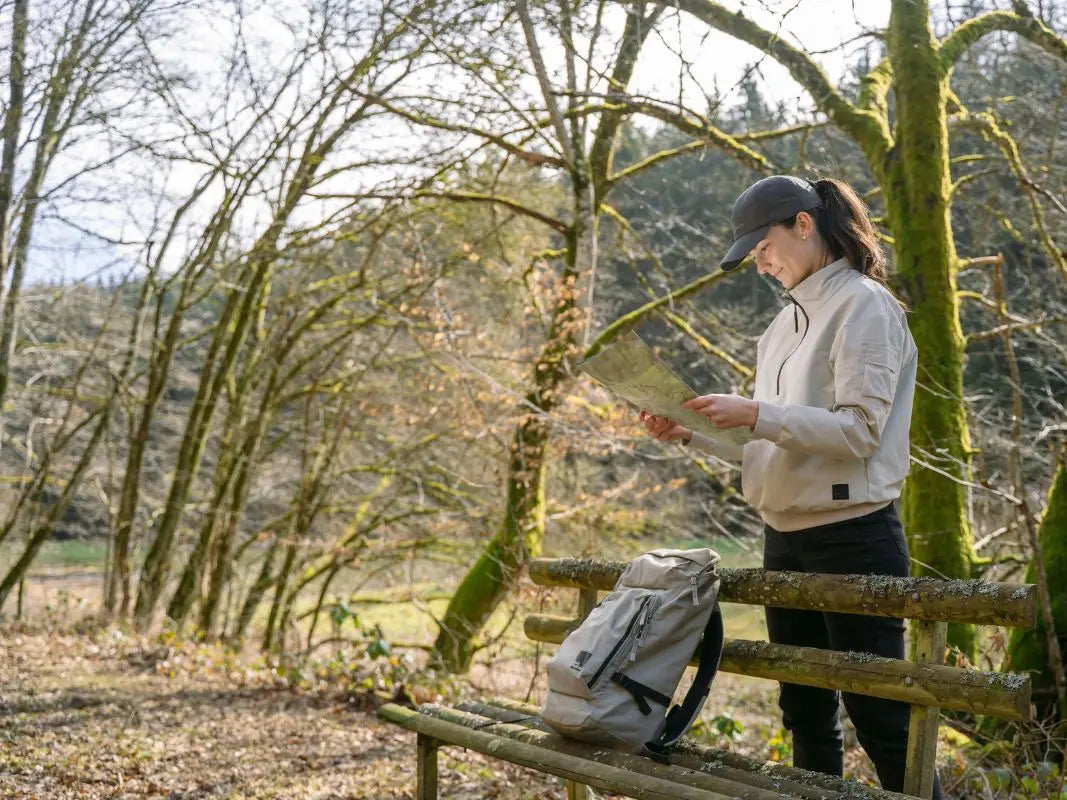 Eine Frau in leichter Jacke liest auf einer sonnigen Waldbank in der Nähe von Wohllebens Waldakademie eine Wanderkarte Vulkaneifel, den Rucksack neben sich.