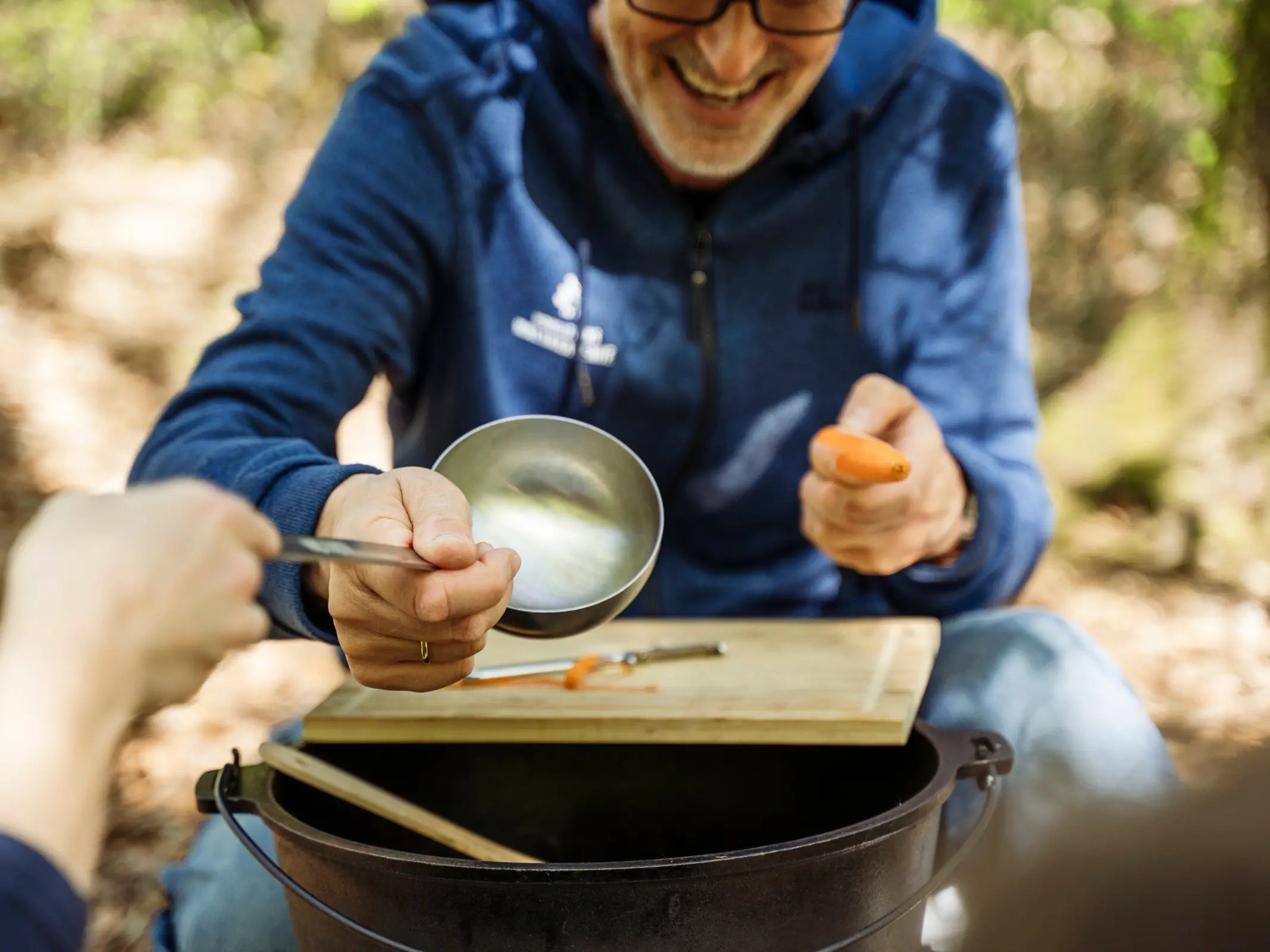 Eine lächelnde Person bereitet während einer Wanderung mit Wohllebens Waldakademie im Freien Essen zu. Sie hält eine Schüssel und eine Karotte neben einem Schneidebrett und einem Topf im Buchenwälder.